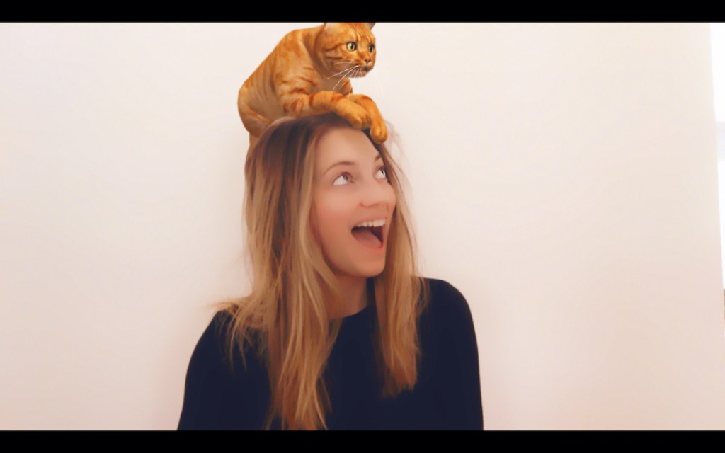 woman sitting against white wall with orange cat on head