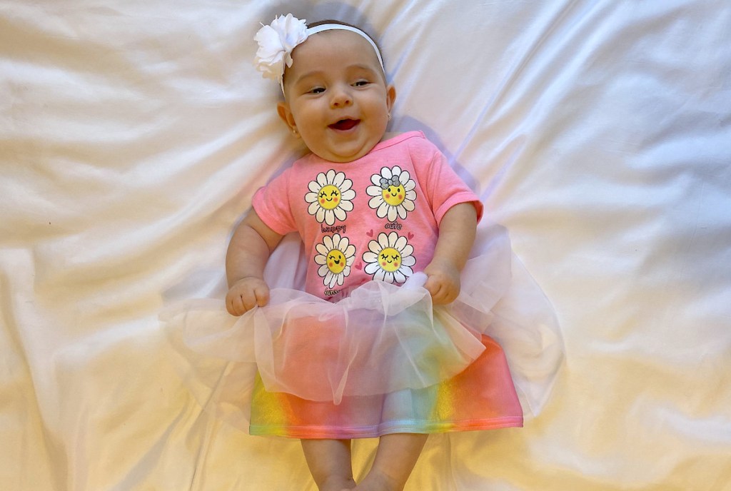 baby girl laying on white blanket with tutu skirt flower shirt and headband