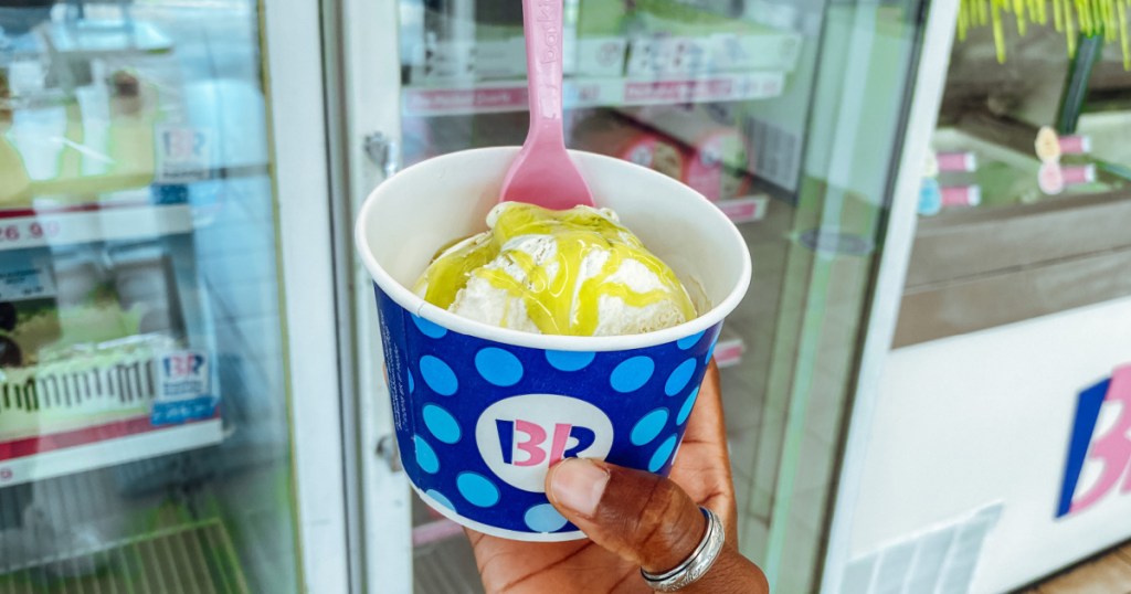 hand holding cup of ice cream and bright green topping and spoon in store