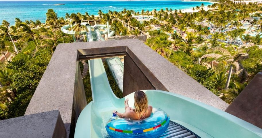 little boy on a round float going down a large waterslide, you can see the beach, ocean, and palm trees in the distance