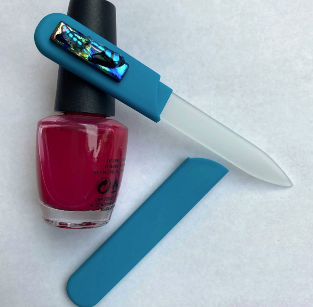 blue and clear glass nail file laying on white surface with bottle of nail polish