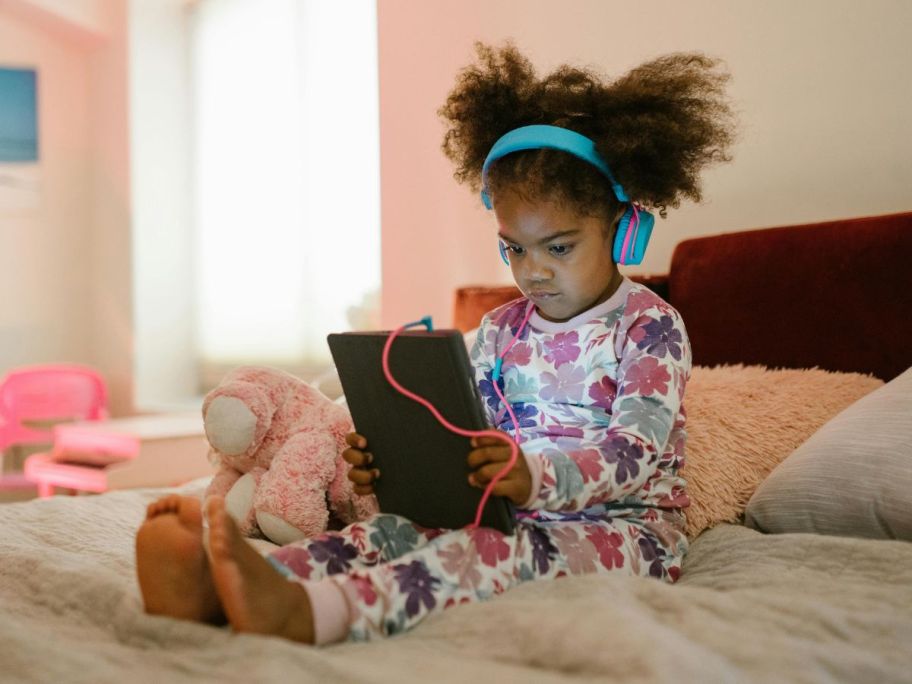 Little girl sitting on a bed watching a tablet while wearing headphones