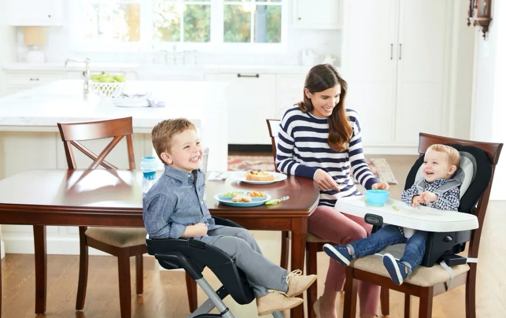 Mom feeding a baby in a highchair and a toddler sitting in a booster seat at a table