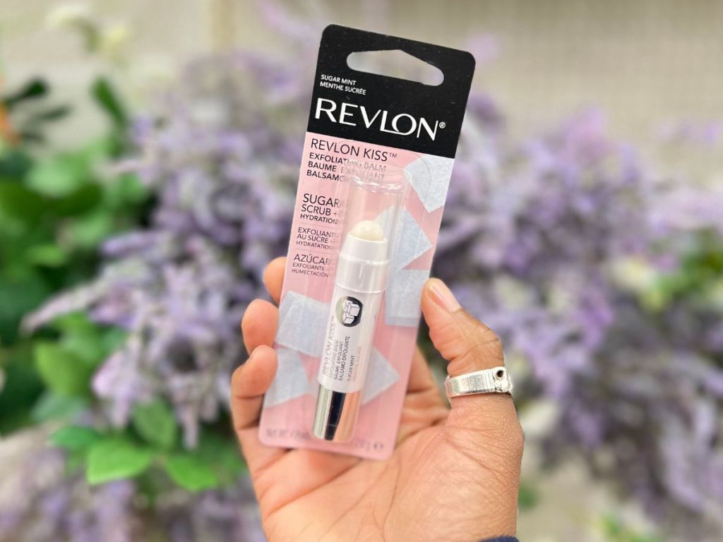 hand holding Revlon Kiss Lip Sugar Scrub Balm in front of purple flowers