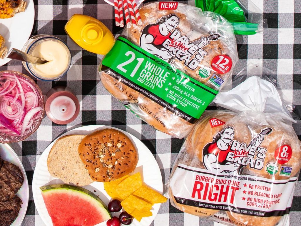 Two packs of Dave's Killer Bread buns on a picnic blanket with picnic foods