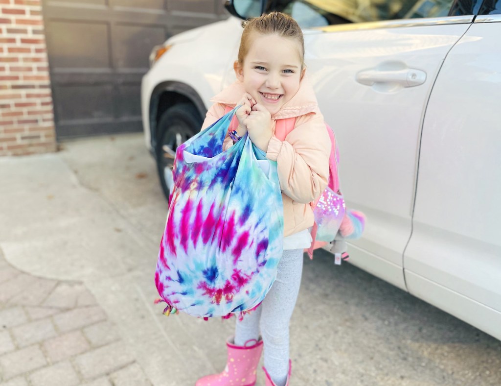 girl smiling holding up tie dye bag next to white car