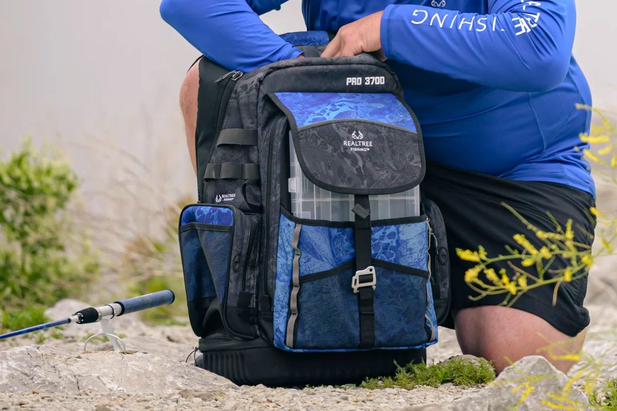 a man kneeling on a lake bank reaching into a blue and black realtree fishing tackle back pack