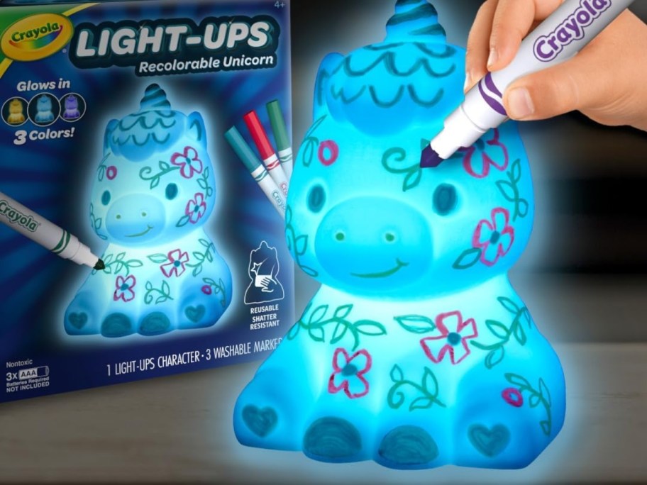 child's hand with a Crayola marker drawing on a a light up unicorn with doodles on it, box this toy onlinees in behind it