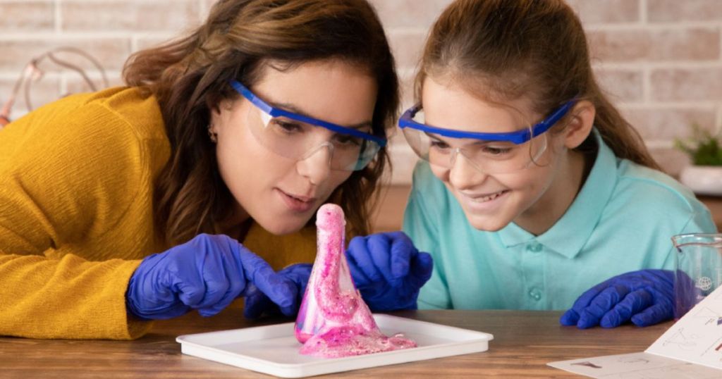 two girls performing a chemistry experiment with a beaker filled with pink foam while wearing safety glasses and protective gloves. 