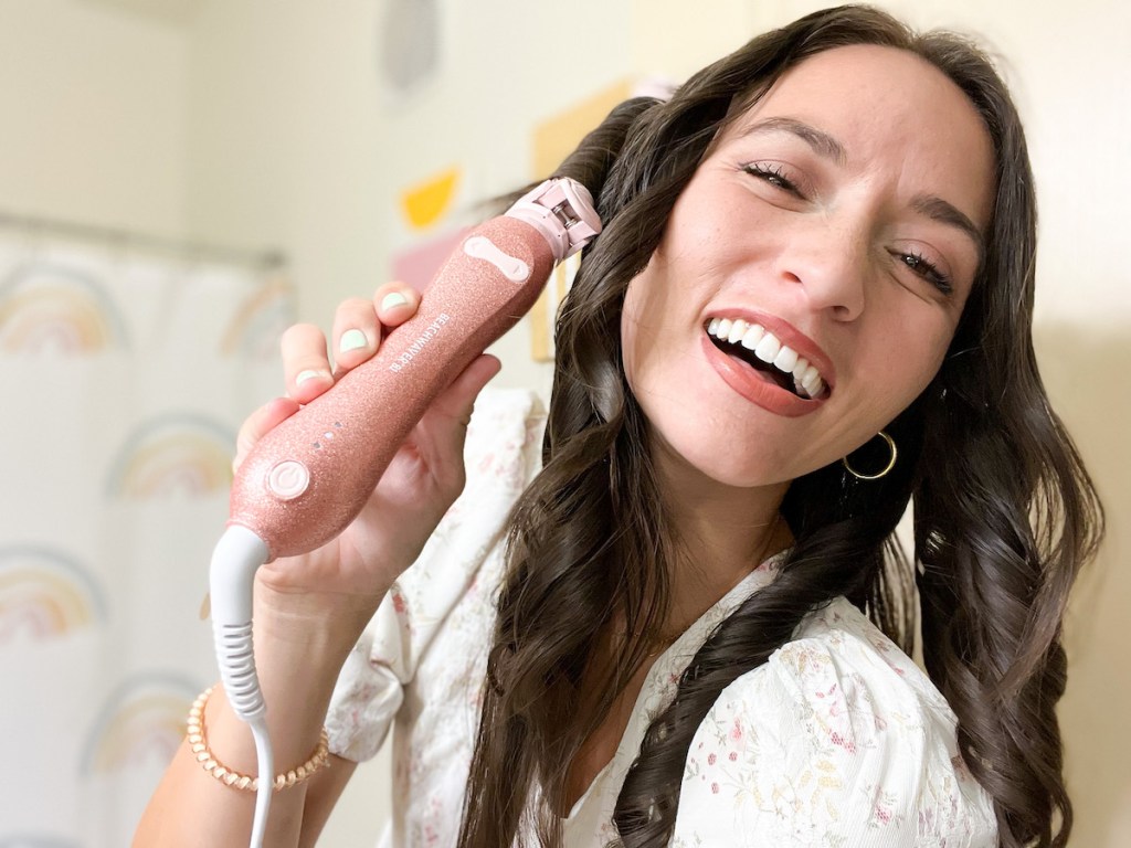 woman curling hair smiling holding pink beachwaver curling wand