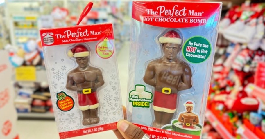 hand holding 2 packs of The Perfect Man chocolate, one is and ornament and one in a hot chocolate bomb, they both look like a man in a Santa Hat and shorts