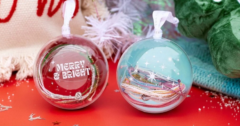 a red and a blue Pura Vida Bracelet Ornament, the ornaments are clear in front and you can see the bracelets inside, Christmas tree and decor behind them