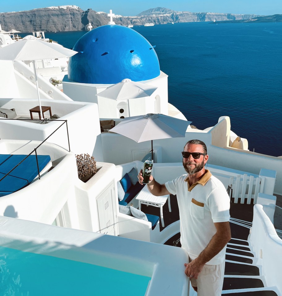 man holding up drink with santorini blue domes and caldrea in background