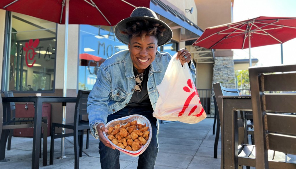 smiling woman holding up a heart tray of chicken nuggets and a to go bag in front of a chick fil a restaurant