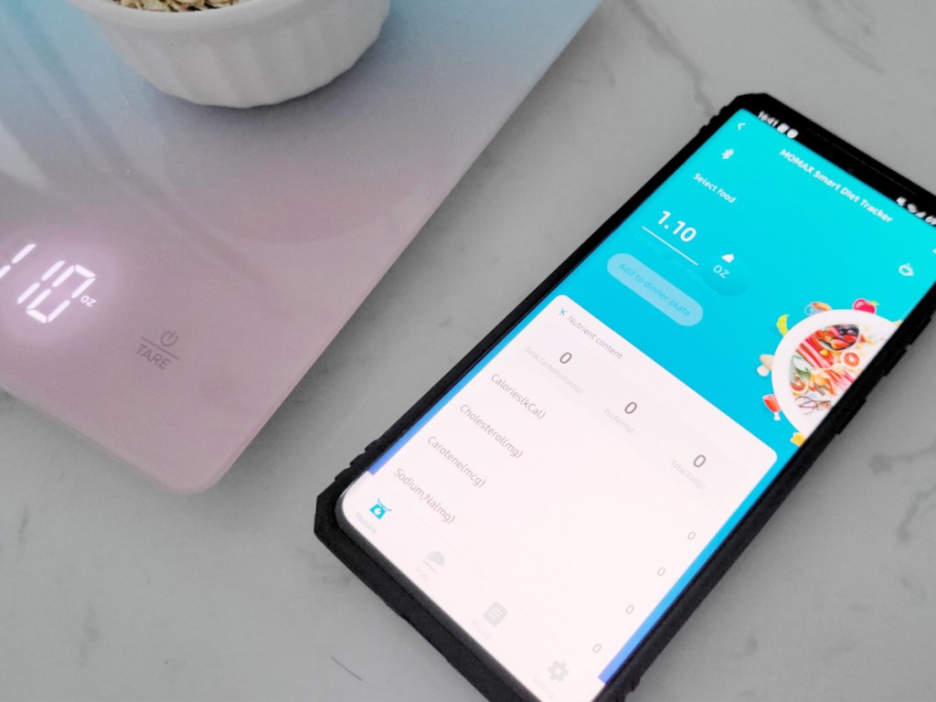 digital scale weighing small bowl of oats next to mobile phone with the Momax Diet app open on screen