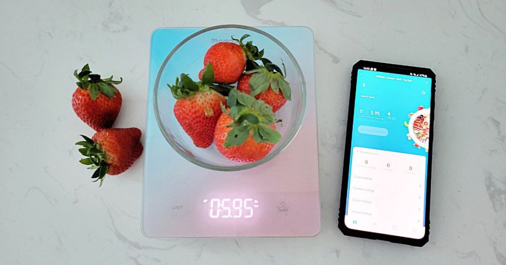 glass bowl full of strawberries on blue and pink glass scale with phone beside it