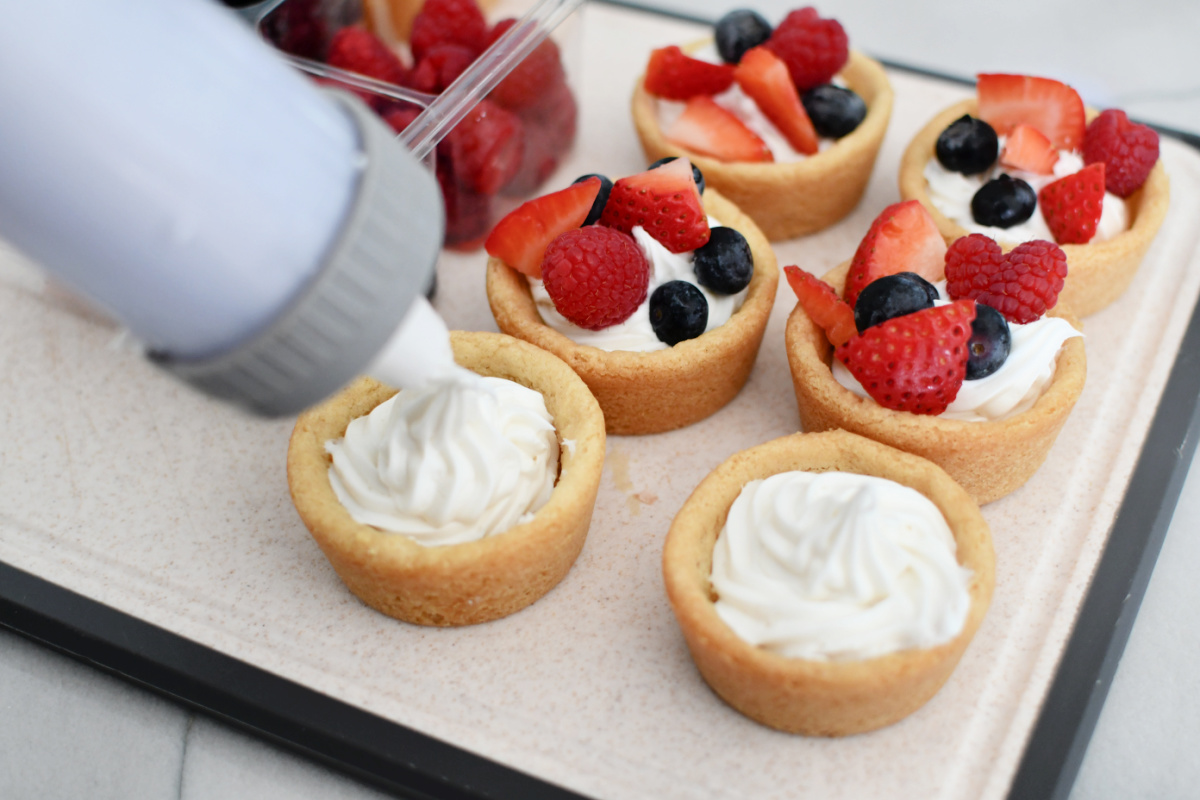 Piping white frosting to sugar cookies with fruit on top. 