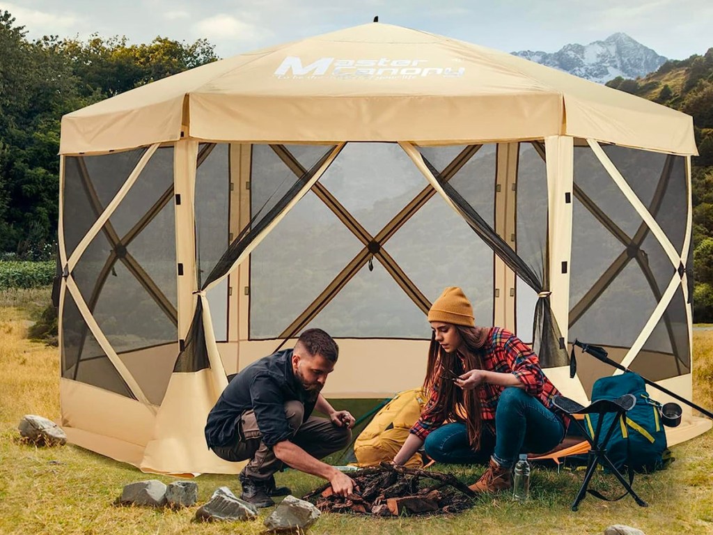 tan mastercanopy shelter with two people building a fire in front of it