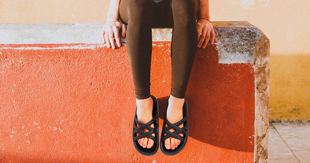 woman sitting on orange wall wearing black sandals and brown leggings