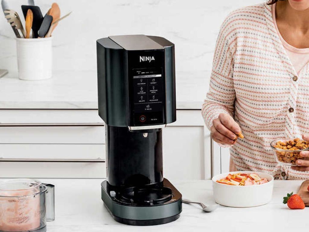 Ninja creami ice cream maker sitting on a counter and a woman adding toppings to ice cream next to it