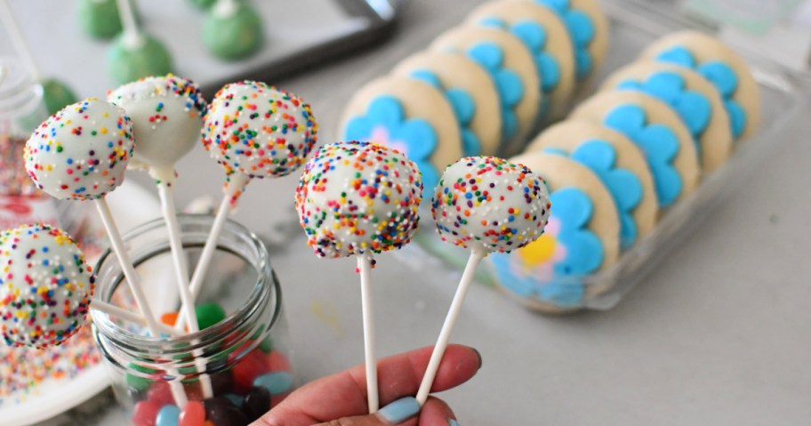 hand holding two cake pops in front of a box of frosted sugar cookies