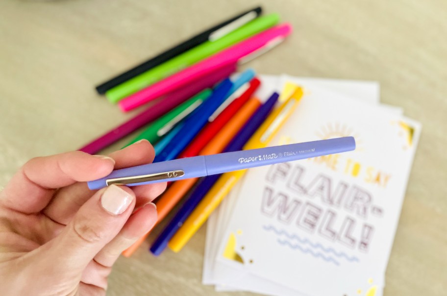 hand holding a light purple flair pen over pile of papermate pens and card