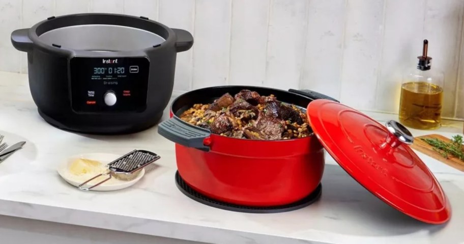 a black Instant Pot Dutch Oven cooker with the red Dutch Oven to the side, lid on the side, with food inside the Dutch Oven on a kitchen counter