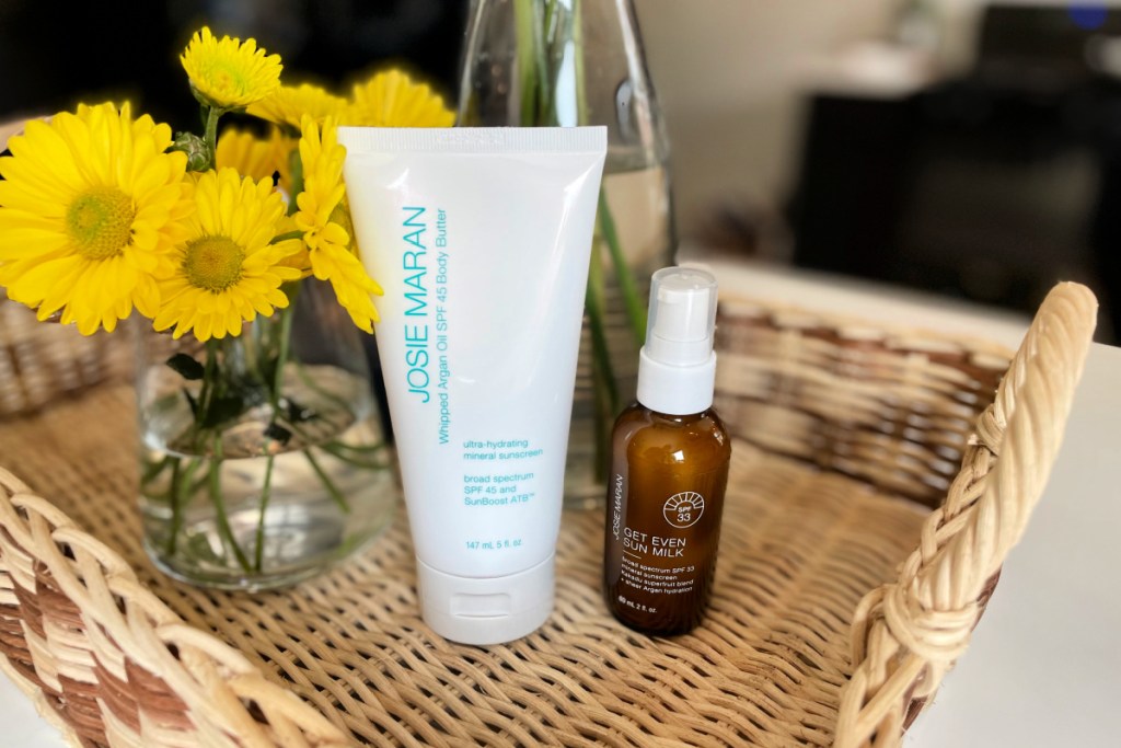 Josie Maran Argan Sun Milk and Body Butter Sunscreen Protection Set in a basket next to yellow flowers