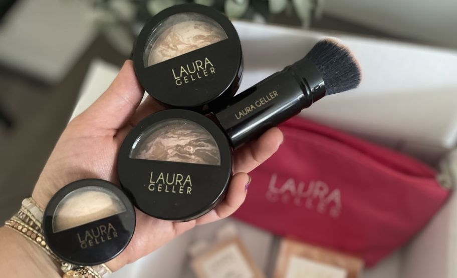 a womans hand holding a baked blush, bronzer, highlighter and brush