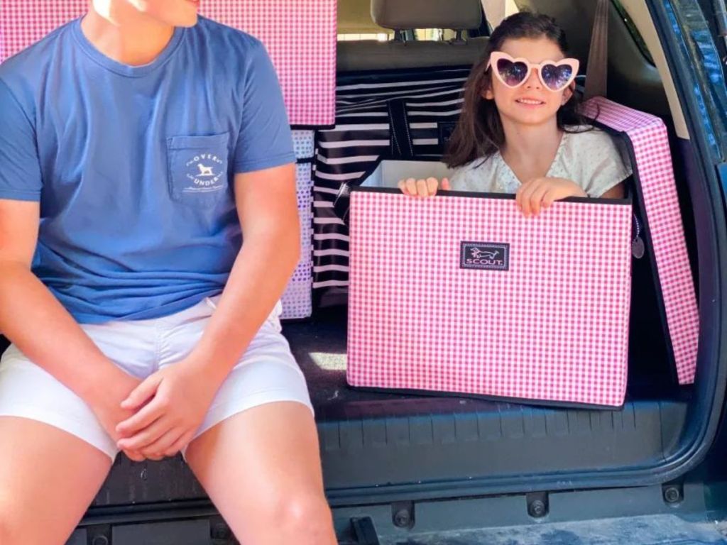 girl sitting in white and pink plaid Scout storage bag in trunk of car