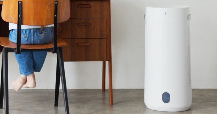 a medium size white air purifier and humidifier in a room, a desk with chair and a little girl sitting in the chair next to it