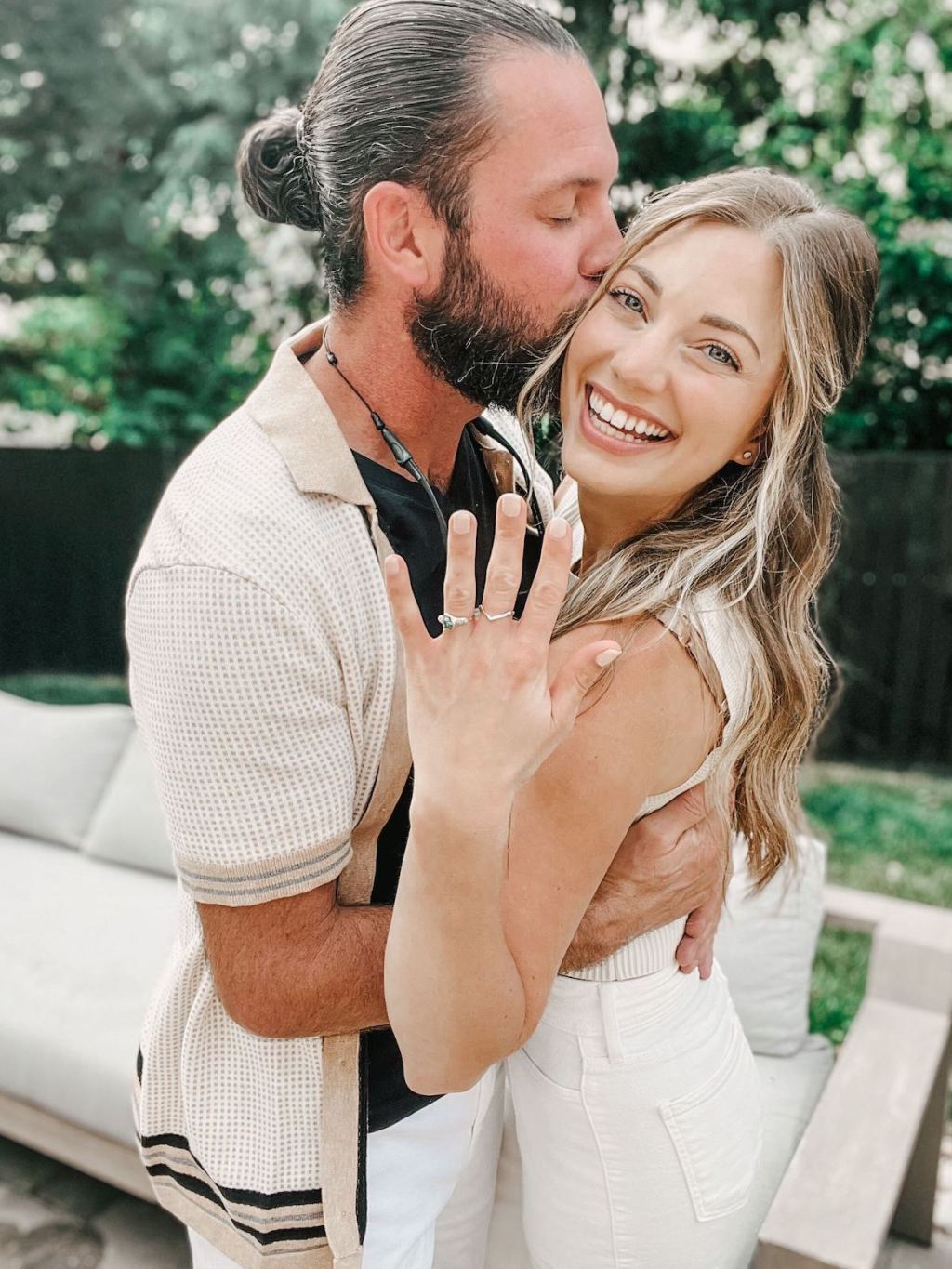 man kissing woman on cheek with woman smiling holding up engagement ring on hand