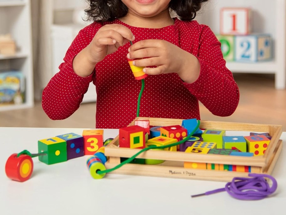 little girl in a red shirt sitting at a table and playing with a set of wooden activity beads and lacing them on a green string