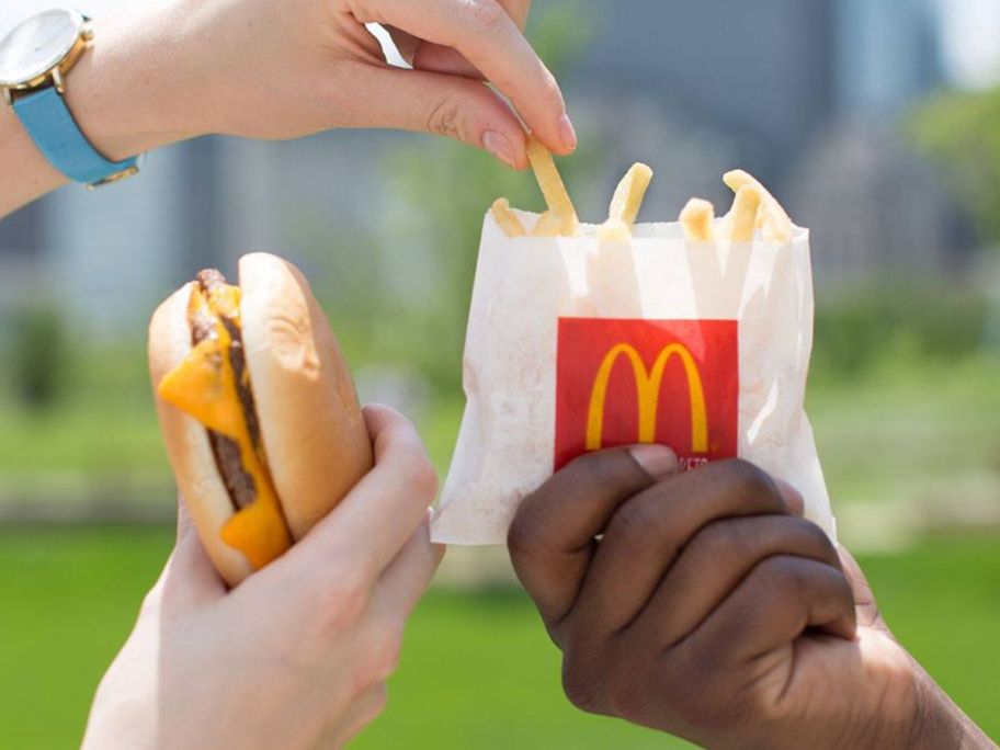 Hand holding a mcdonald's double cheeseburger while taking a french fry from a bag held by another hand