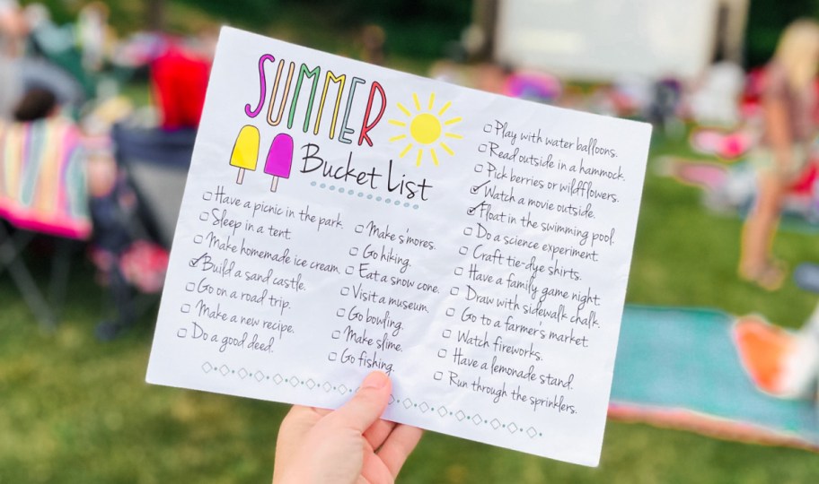 hand holding up a free printable summer bucket list in front of an outdoor movie screen