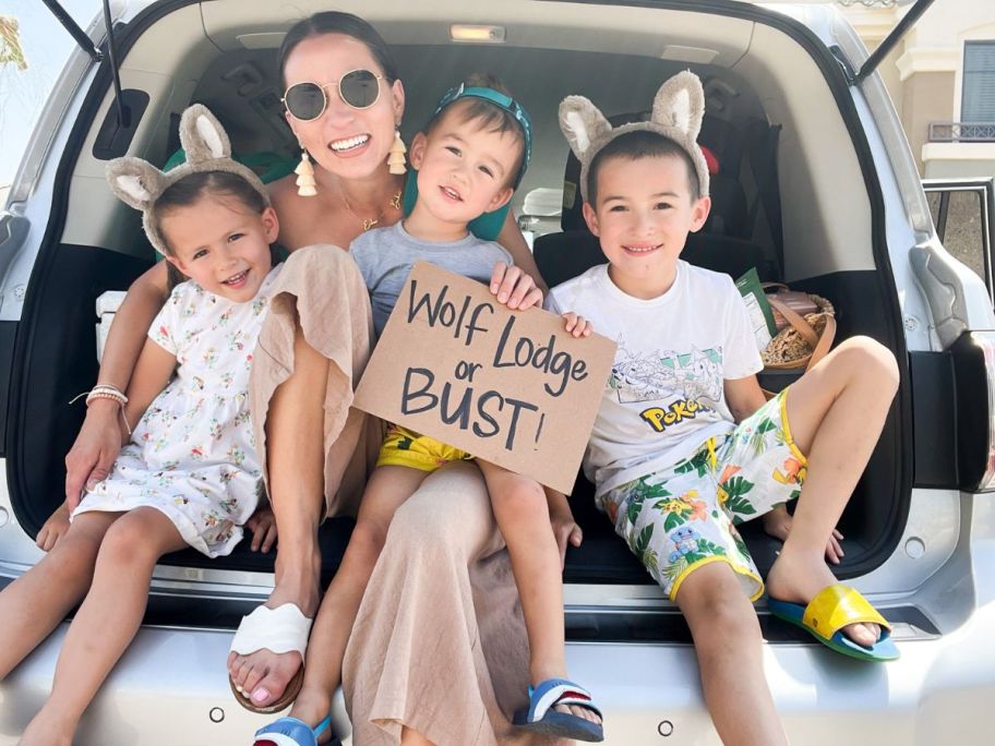 A mom and 3 kids sitting in the back of a vehicle with a Wolf Lodge or Bust! sign