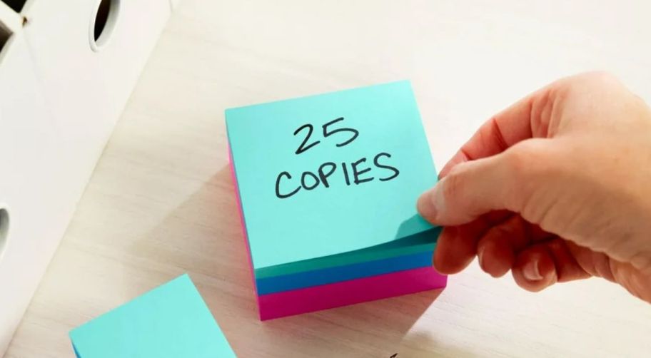 a hand taking the top sheet off a 400 count cube pack of brightly colored post it notes