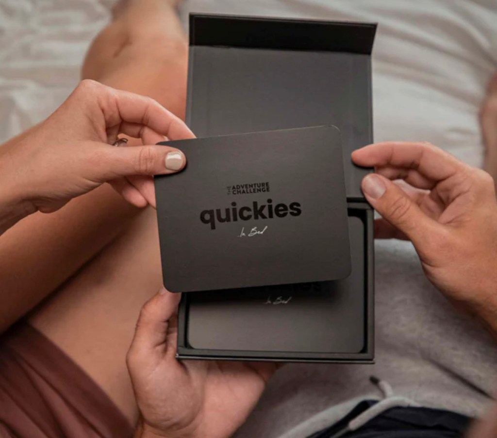 couple sitting on bed holding black quickies scratch off card and box