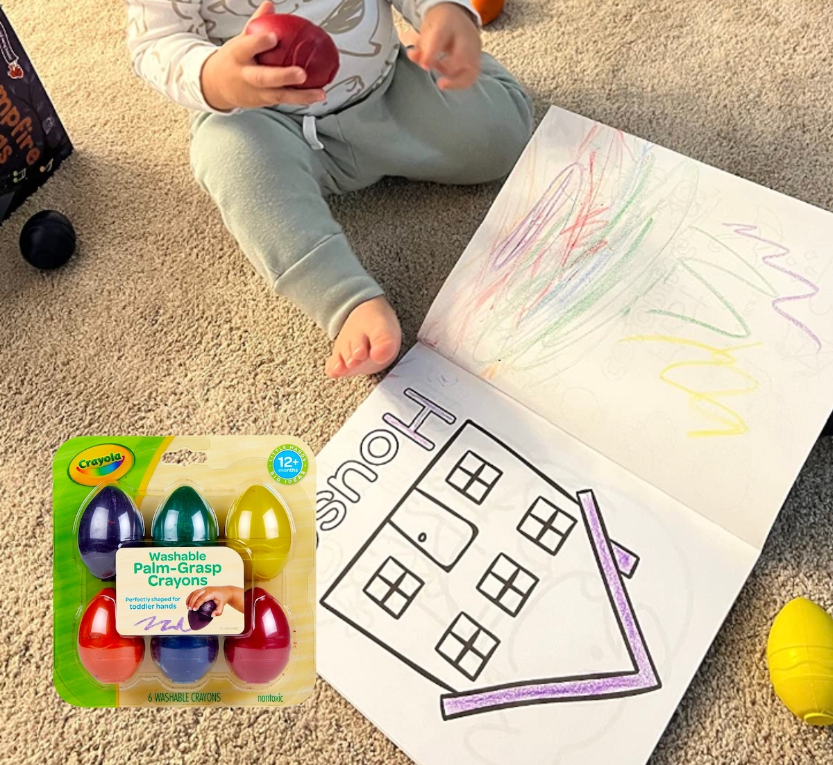 toddler holding red egg shaped crayon sitting in front of house coloring book