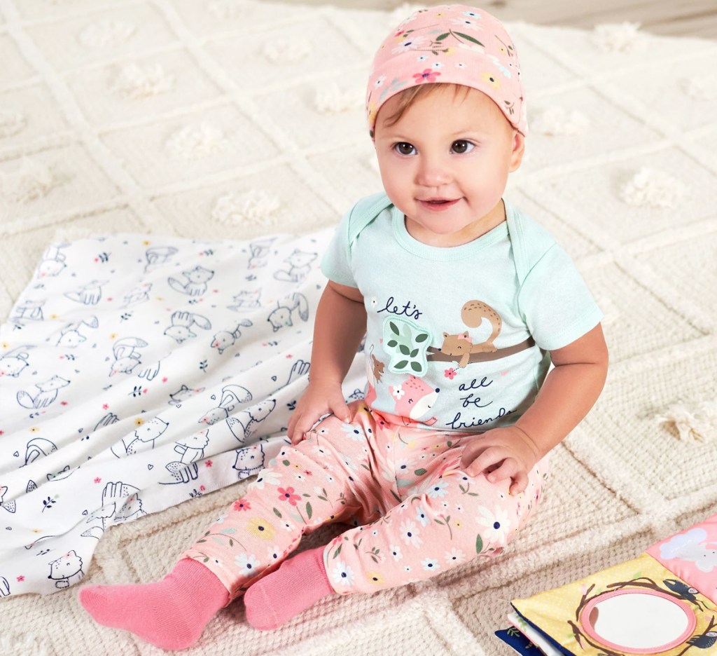 baby sitting on blanket wearing blue onesie, pink pants, and matching hat