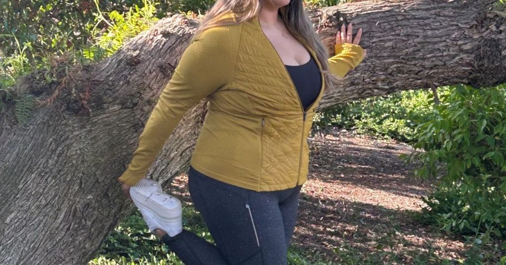 woman wearing a mustard color Athleta jacket stretching during a workout / walk outside