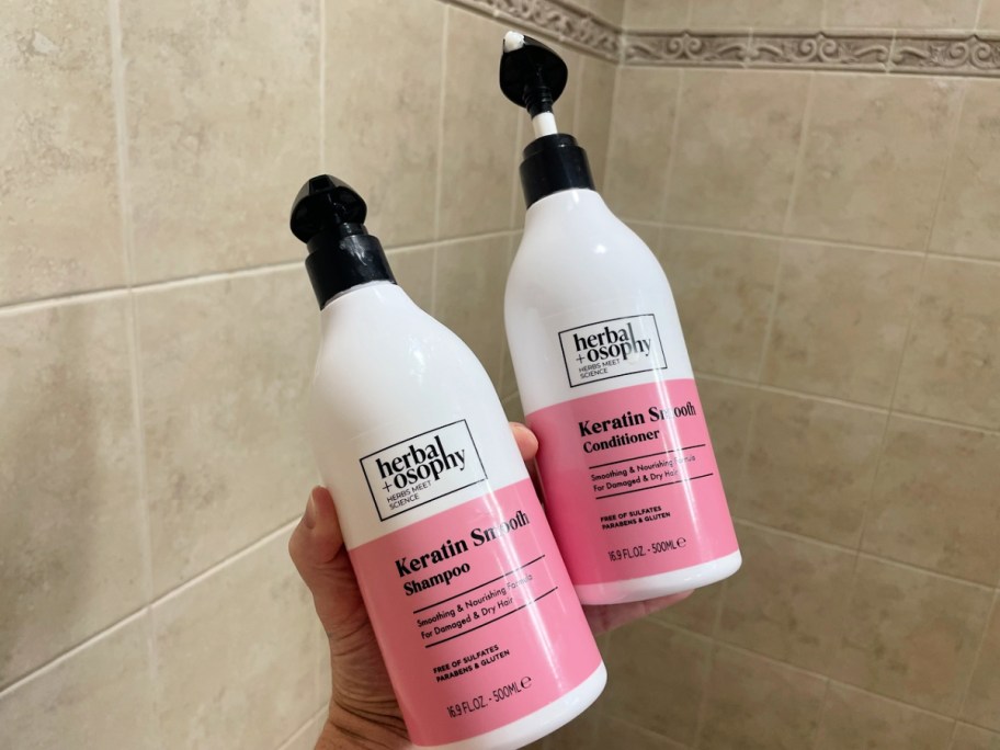 pink and white Herbalosophy Keratin Shampoo and Conditioner Set in hand with shower tile background