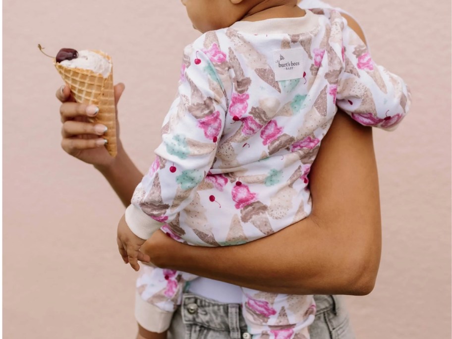 mom holding a toddler, toddler's back to the camera, wearing a pajama set with ice cream cones on it, mom is holding a real ice cream cone