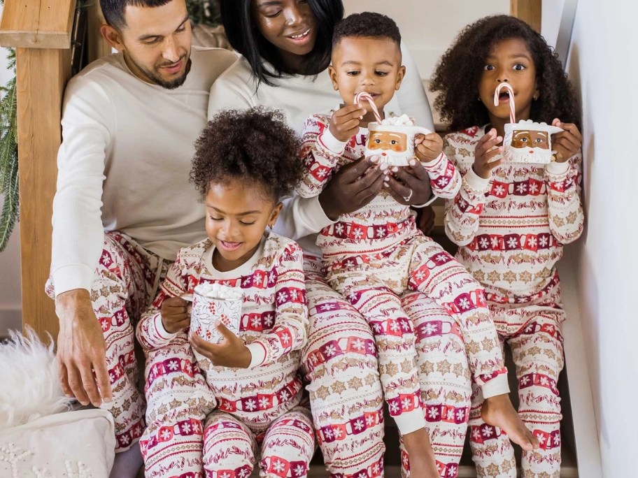 family sitting on stairs wearing matching holiday pajamas that have hot chocolate on them