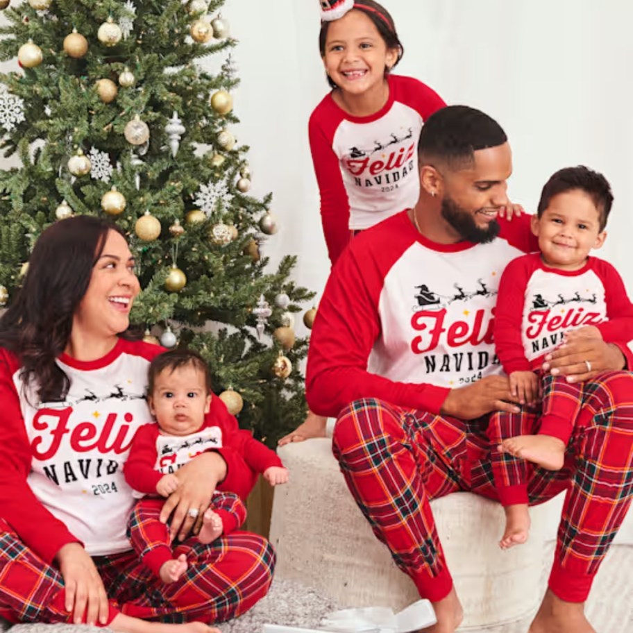 family by a Christmas tree wearing matching red/black plaid and white long sleeve Christmas pajamas that say "Feliz Navidad"