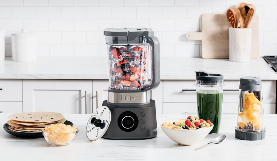 a ninja foodi blender and two blender cups, a blending disc, a plate of tortillas, a small glass bowl of shredded cheese, a smoothie bowl, and a spoon on a white marble kitchen counter.