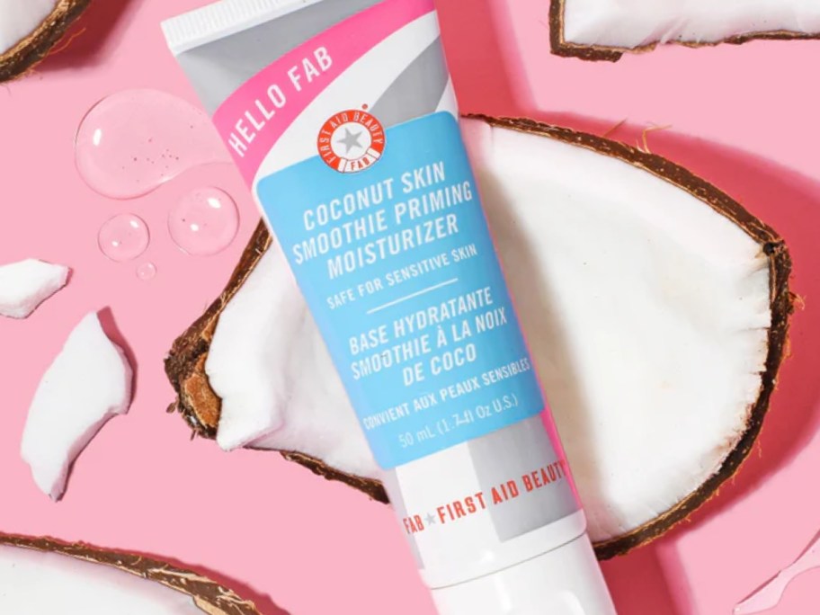 tube of First Aid Beauty Hello FAB Coconut Skin Smoothie Priming Moisturizer on top of pieces of coconut on a pink background