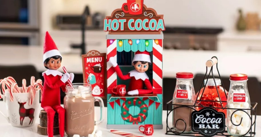 Elf on The Shelf dolls on a kitchen counter with a prop Hot Cocoa stand and hot cocoa and extras around them