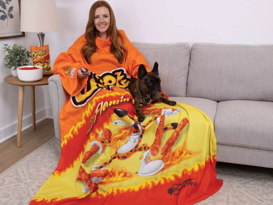 Woman sitting on a couch with a dog while wearing a Flamin' Hot Cheetos Snuggie