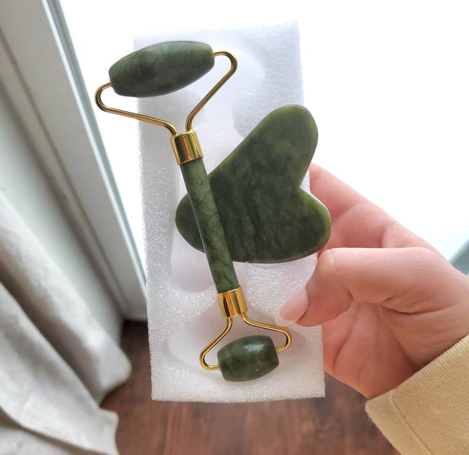 hand holding jade roller and gua sha facial tools by window